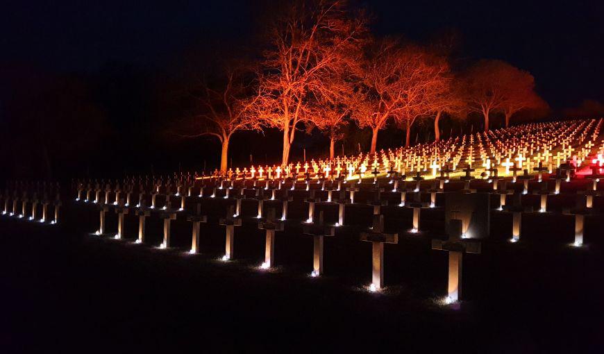 Cimetière militaire français de Craonnelle illuminé de nuit, avec des rangées de croix éclairées par des bougies et des arbres teintés de lumière rouge en arrière-plan.