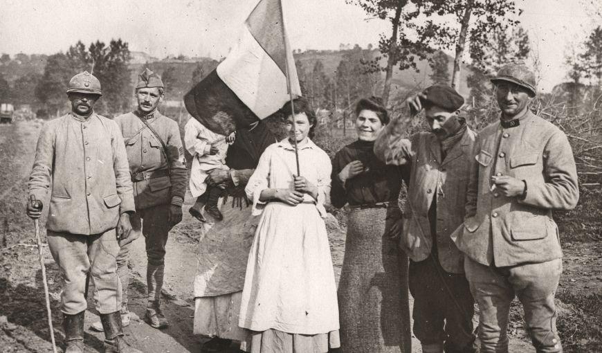 Groupe de soldats français et de civils posant ensemble sur un chemin rural pendant la Première Guerre mondiale, dont une femme tenant un drapeau tricolore, avec un paysage vallonné en arrière‑plan.