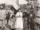 Groupe de soldats français et de civils posant ensemble sur un chemin rural pendant la Première Guerre mondiale, dont une femme tenant un drapeau tricolore, avec un paysage vallonné en arrière‑plan.