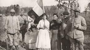 Groupe de soldats français et de civils posant ensemble sur un chemin rural pendant la Première Guerre mondiale, dont une femme tenant un drapeau tricolore, avec un paysage vallonné en arrière‑plan.
