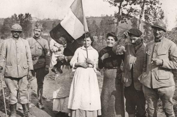 Groupe de soldats français et de civils posant ensemble sur un chemin rural pendant la Première Guerre mondiale, dont une femme tenant un drapeau tricolore, avec un paysage vallonné en arrière‑plan.
